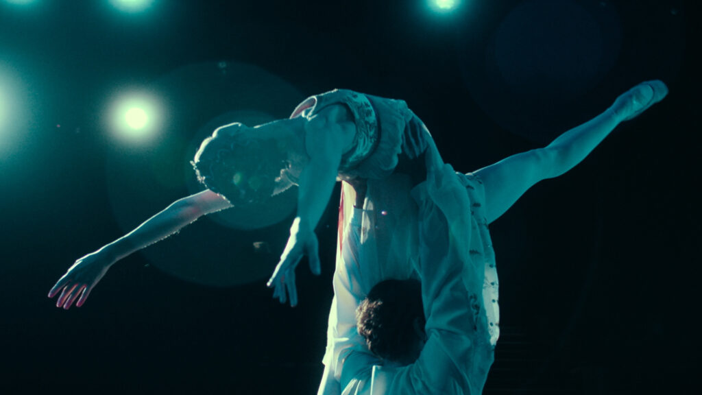 A close-up from behind of two dancers in a pas de deux. One dancer holds the other overhead; the partner extends one leg devant and arches back, arms in a V, over the supporting partner's head.