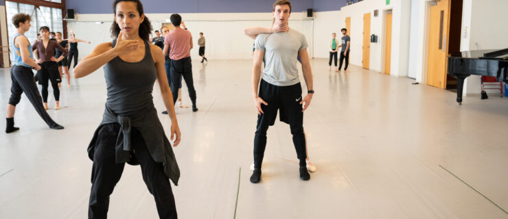 Claudia Schreier, in black athletic clothing, stands wide-legged at the front of a large rehearsal studio, one arm dropped as the other lifts up and bends at the elbow as if gripping something in front of her. She speaks to a pair of dancers behind her. The frontmost dancer stands with his arms down as the one behind him mimics Schreier's position, her hand around her partner's throat.