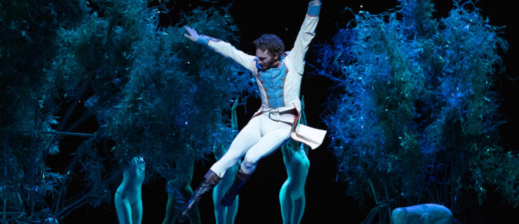 Skylar Campbell, wearing a white jacket with tails, white tights, and black boots, jumps up and tucks his legs slightly. He lifts his arms up and looks down towards his feet. Behind him, dancers in unitards stand behind trees.