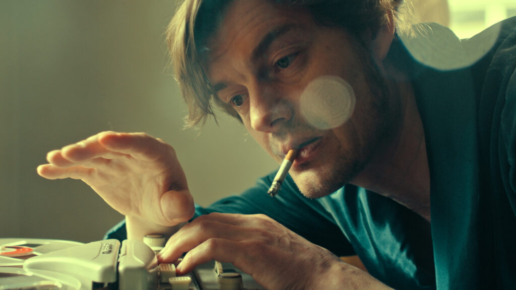 An actor portraying John Cranko leans over his desk, a cigarette hanging out of his mouth, as he lifts his hands to mark choreography. He has an intense yet dreamy expression in his eyes.