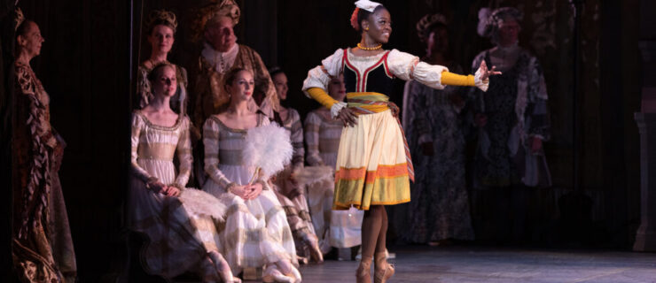 Michaela Mabinty DePrince poses in sous-sus onstage with her right hand on her hip and her left arm extended out to the side. She looks over her left should and smiles. She wears a multi-colored, knee-length dress, brown pointe shoes and a red and white headpiece. Other dancers in aristocratic, medieval costumes sit on stools behind her and watch.