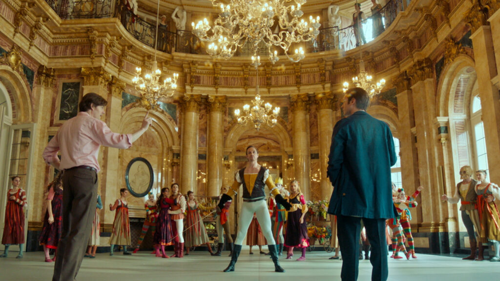 In a grand, ornate ballroom with gold accents and several chandeliers, a group of dancers rehearse a scene from "Romeo and Juliet." Two men stand at the front, talking to each other as they direct the rehearsal. One man stands in costume, holding a prop sword, in the middle of the floor.