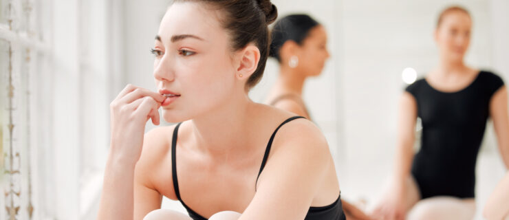 A teenage ballet dancer is shown sitting on the floor from the waist up. She pulls her knees up to her chest and wraps her arms around them, and bites her fingernail nervously as she looks out the dance studio window. Behind her, two female dancers in black leotards and pink tights sit on the floor and talk.