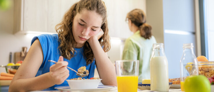 Upset and distraught teen girl eating breakfast in the kitchen while her mother is doing chores