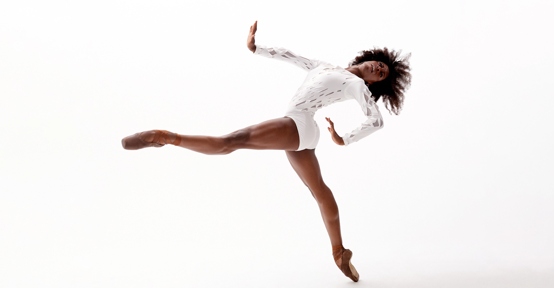 April Watson, wearing a long-sleeved white leotard, stands on pointe and does a 90 degree battement with her left leg as she leans back with her upper body. She pushes throws her head back and pushes her right arm out, her left arm bent and closer to her side.