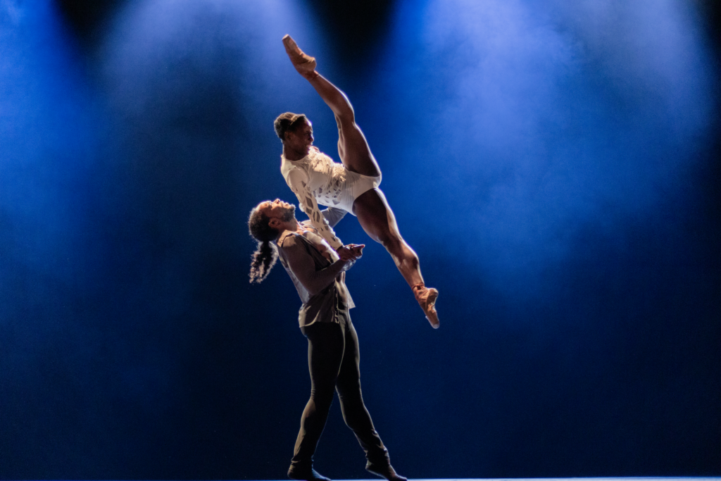 A male dancer lifts a ballerina dramatically up into the air, holding her right hand and back as she splits her legs. She wears a white leotard, while he wears gray tights and shirt, and they dance in front of a blue backdrop.