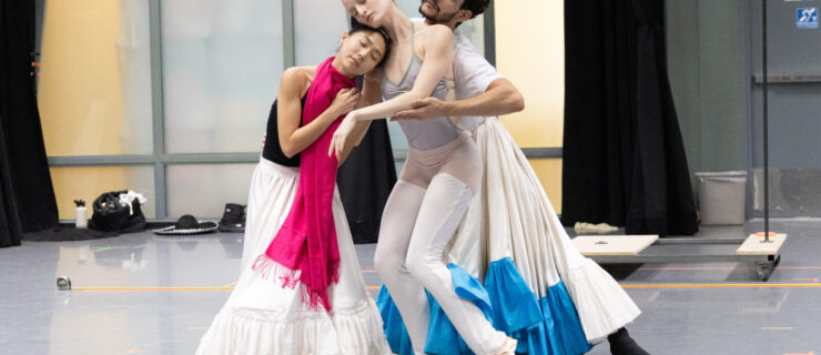 Three dancers rehearse in a ballet studio. They pose together, gently leaning on each other, as the most front and furthest behind dancers wear long, ruffled Mexican-style skirts.