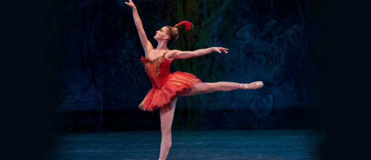 Ashley Bouder, as the Firebird, does a piqué arabesque on pointe onstage with arms in high arabesque. She looks up past her fingertips and wears a bright red, short tutu with gold accents and a red feather headpiece with a gold base.