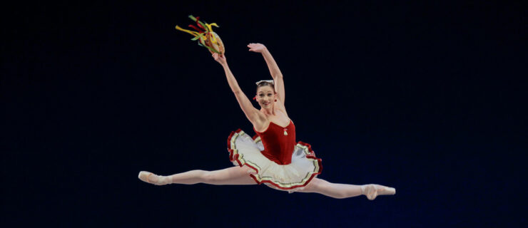 Ashley Bouder, wearing a tutu with a red velvet bodice and white ribbon-lined skirt, is caught at the height of a huge saut de chat, her face beaming. The ribbons on the tambourine that she holds in her right hand stream out behind her.