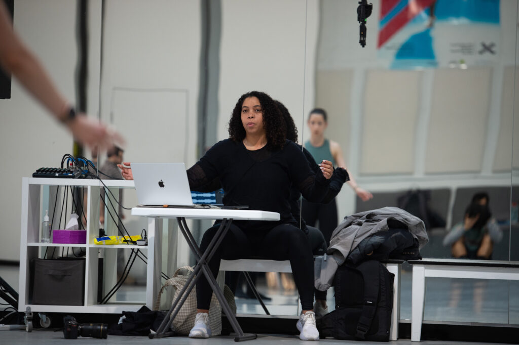 Jennifer Archibald sits on a chair, back to the mirror, in a large dance studio. Her laptop is open on a table in front of her, and she watches the dancers.