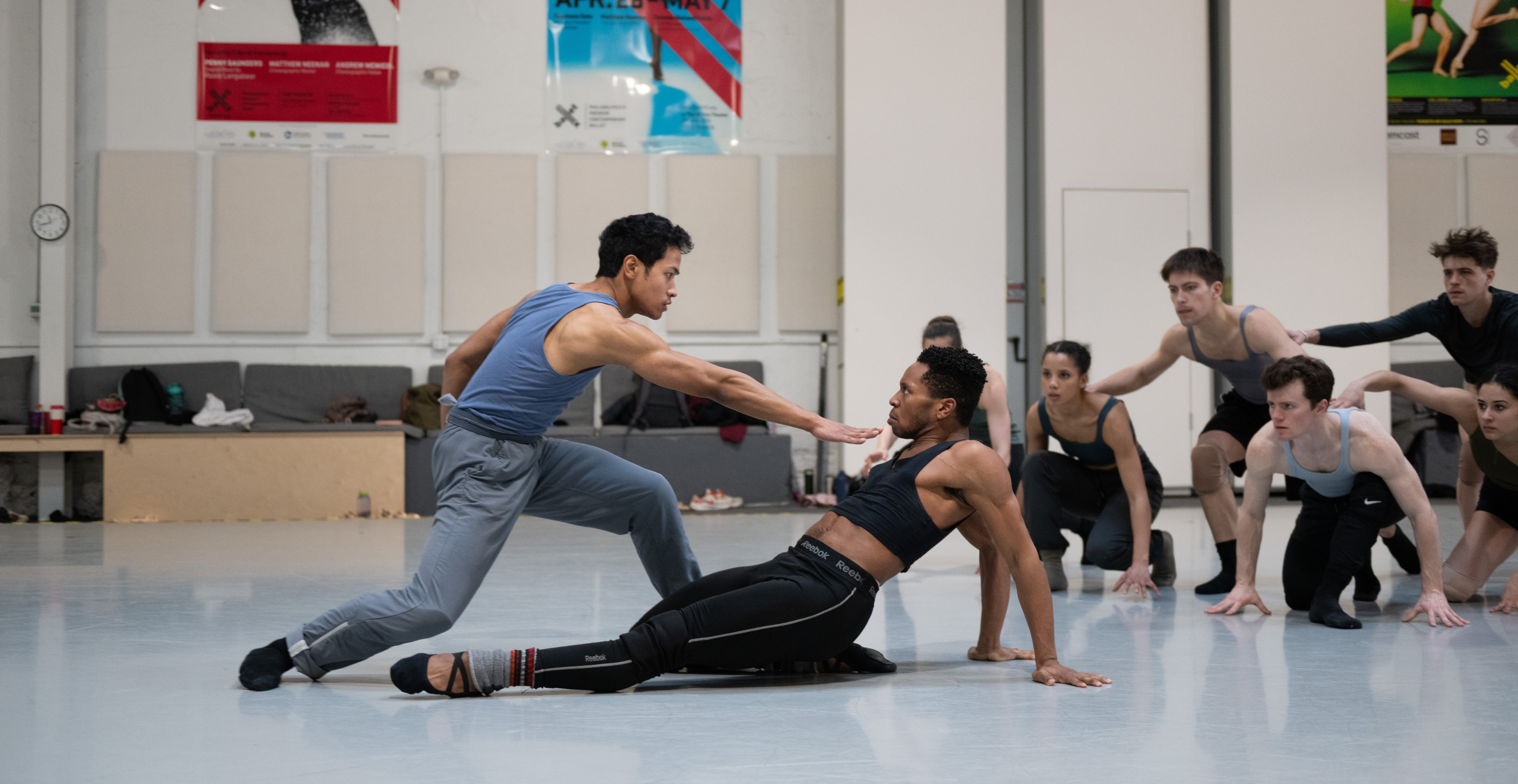 In a large dance studio, two dancers rehearse. One dancer leans back, arms propping up his upper body, with one leg extended front and the other tucked under. The other dancer does a deep lunge with one leg in front as he reaches forward with one arm, hand splayed right in front of his partner's chest. They look into each other's eyes intensely. In the background, a group of dancers crouch with their hands on the ground and watch the duo.