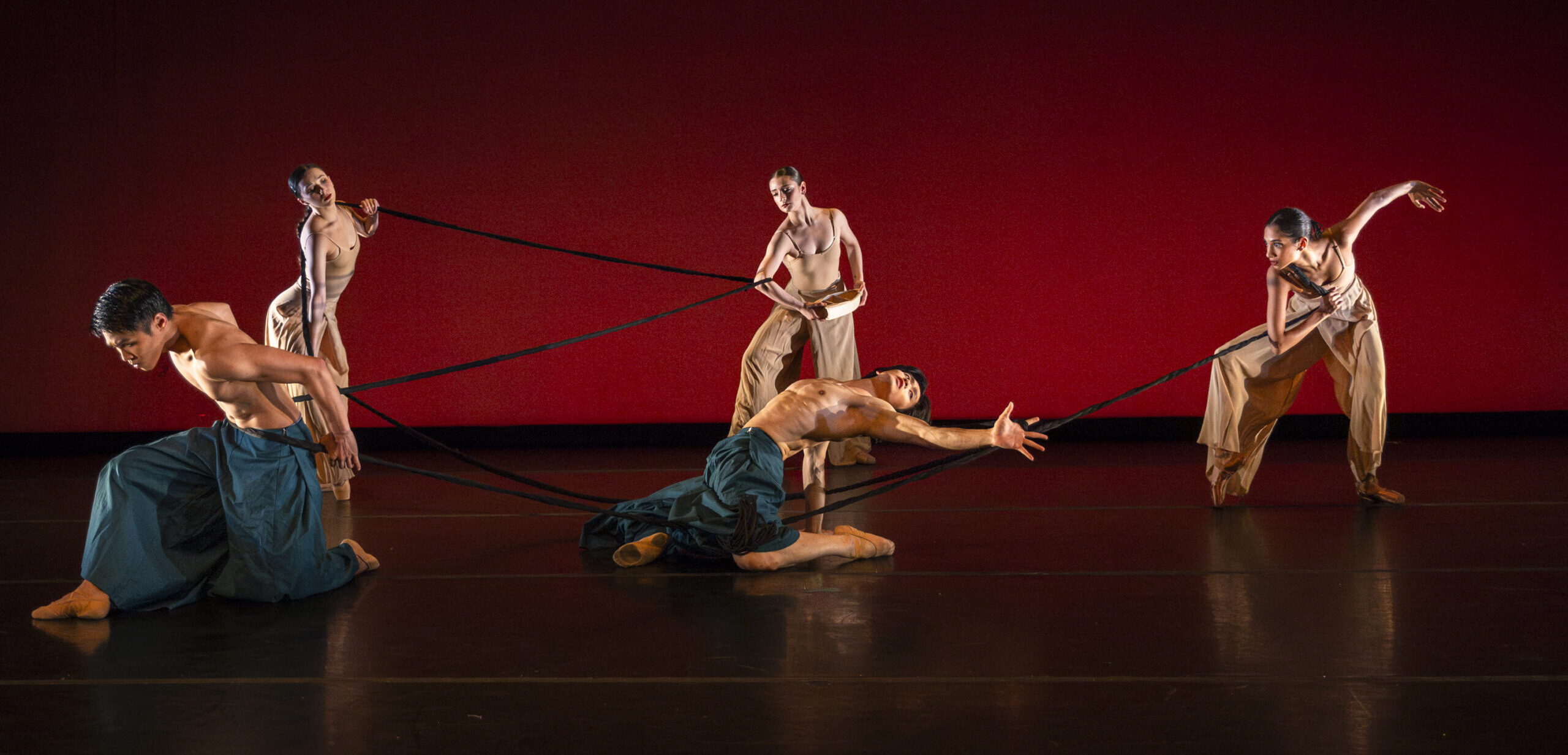 Five Oakland Ballet dancers perform onstage, each holding onto a long black rope. Three dancers wear flowing costumes in tan, and the other two wear billowing teal pants, contrasting with a bold red background. Their facial expressions range from intense focus to serene determination, and they bend forward or arch back, legs bent.