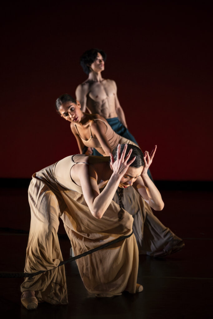 Three Oakland Ballet dancers dance onstage in a vertical line. In the foreground, a dancer in a flowing beige costume arches forward, pressing her hands to her head, with an intense expression of concentration. Behind her another dancer bends forward with a calm, neutral expression. The third dancer, positioned higher and slightly behind, stands erect and looks up and outward.