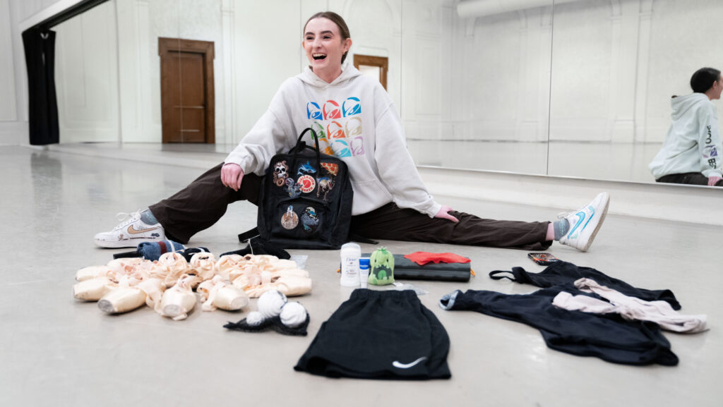 Maya Schonbrun, a fair-skinned dancer with brown hair in a bun, sits on a gray marley floor in front of a mirror. She wears and gray Taco Bell sweatshirt and black sweats. The contents of her dance bag are laid in front of her.