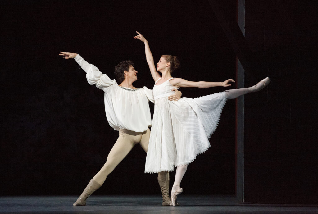 Guillaume Côté partners Elena Lobsanova onstage, who stands in arabesque on pointe with her arms in high third. She looks lovingly at Côté, who lunges in parallel toward her with one arm around her waist and the other extended high. He wears a white billowing tunic and tan tights and boots, and she wears a long white dress.