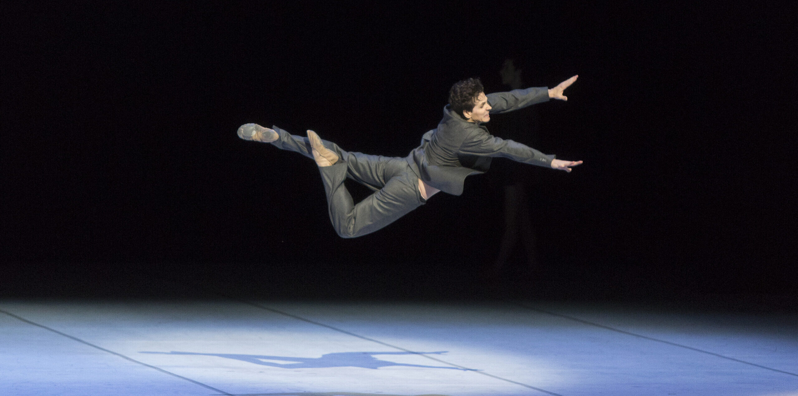 Onstage with a black background and white floor with spotlights, Guillaume Côté flies through the air, stomach facing down, with one leg tucked in passé as the other extends back. He reaches forward with both arms, looking forward with a strained expression. He wears a gray suit.