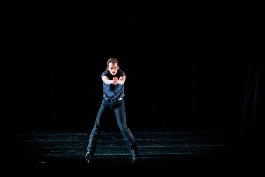 On a very dark stage, Jonathan Harris stands in parallel with legs slightly bent, arms clasped firmly in front as he looks past them toward the audience. He wears black jeans with a belt, and a black shirt.