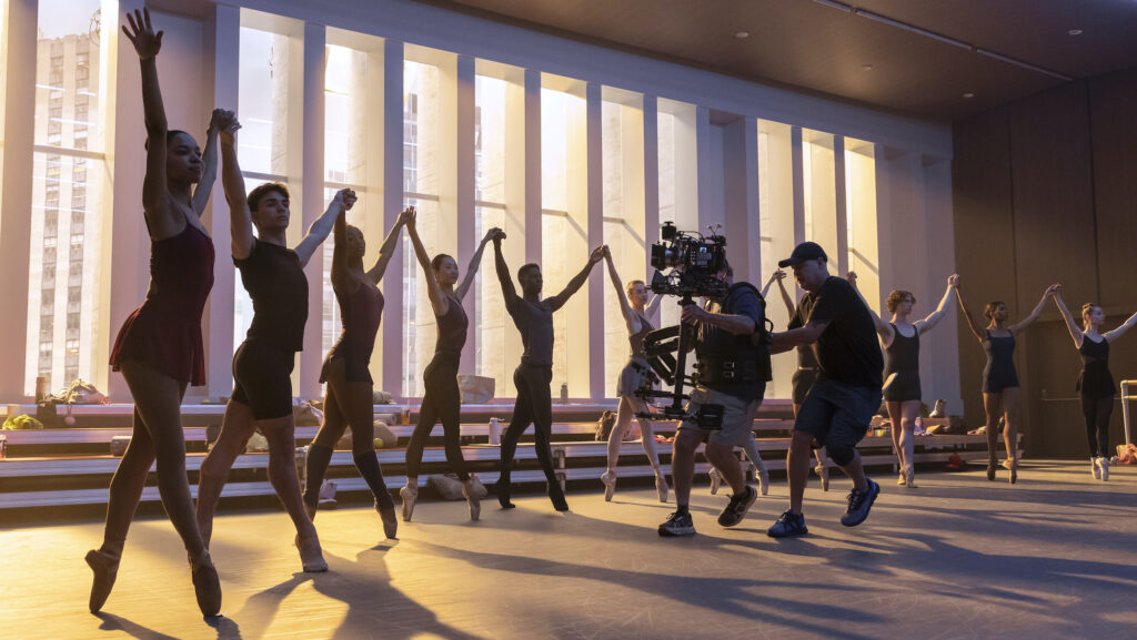 In a dance studio lined with long, narrow windows, a line of male and female dancers stand in fourth position relevé, with the women on pointe, holding their arms up in a V-shape and joining hands. A team of cameramen run by them with a camera, shooting the scene.