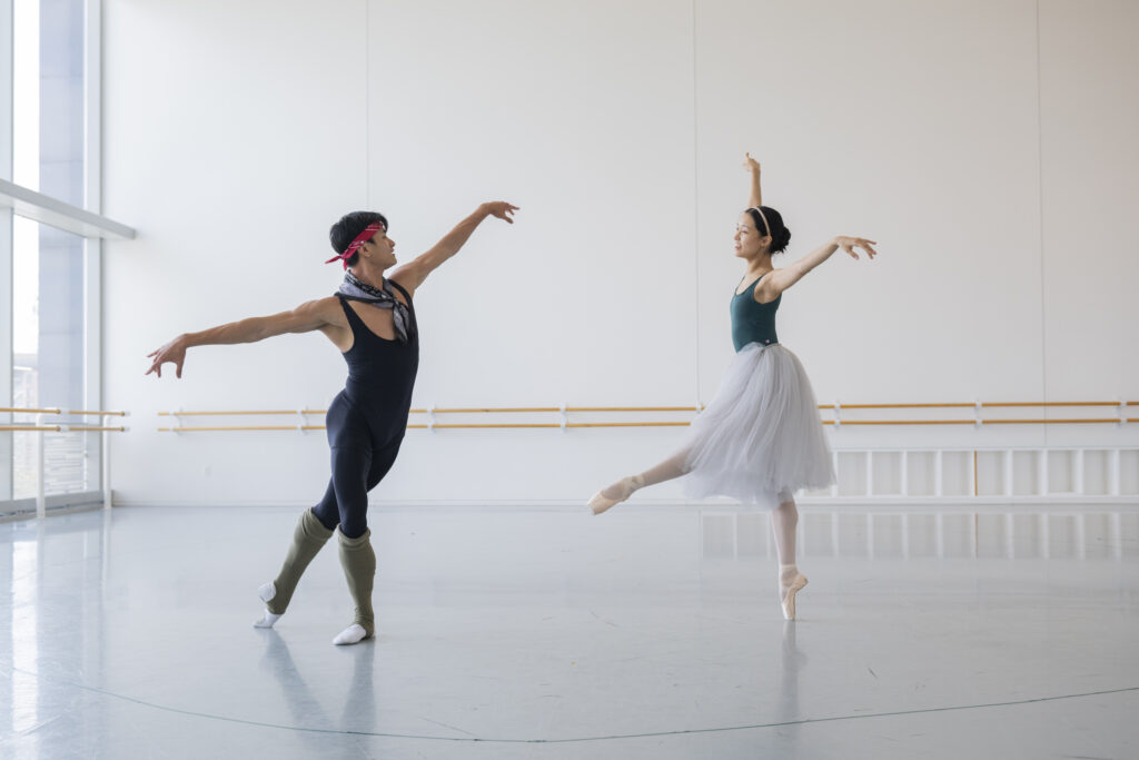 In a bright, airy dance studio, Chun Wai Chan and Danbi Kim rehearse a pas de deux. Chan stands in tendu derriere with a slight fondu, arms open and long. Kim is mid-movement, poised on one pointe with her other leg extended in front on the croisé devant line, arms raised in a gentle “V.”