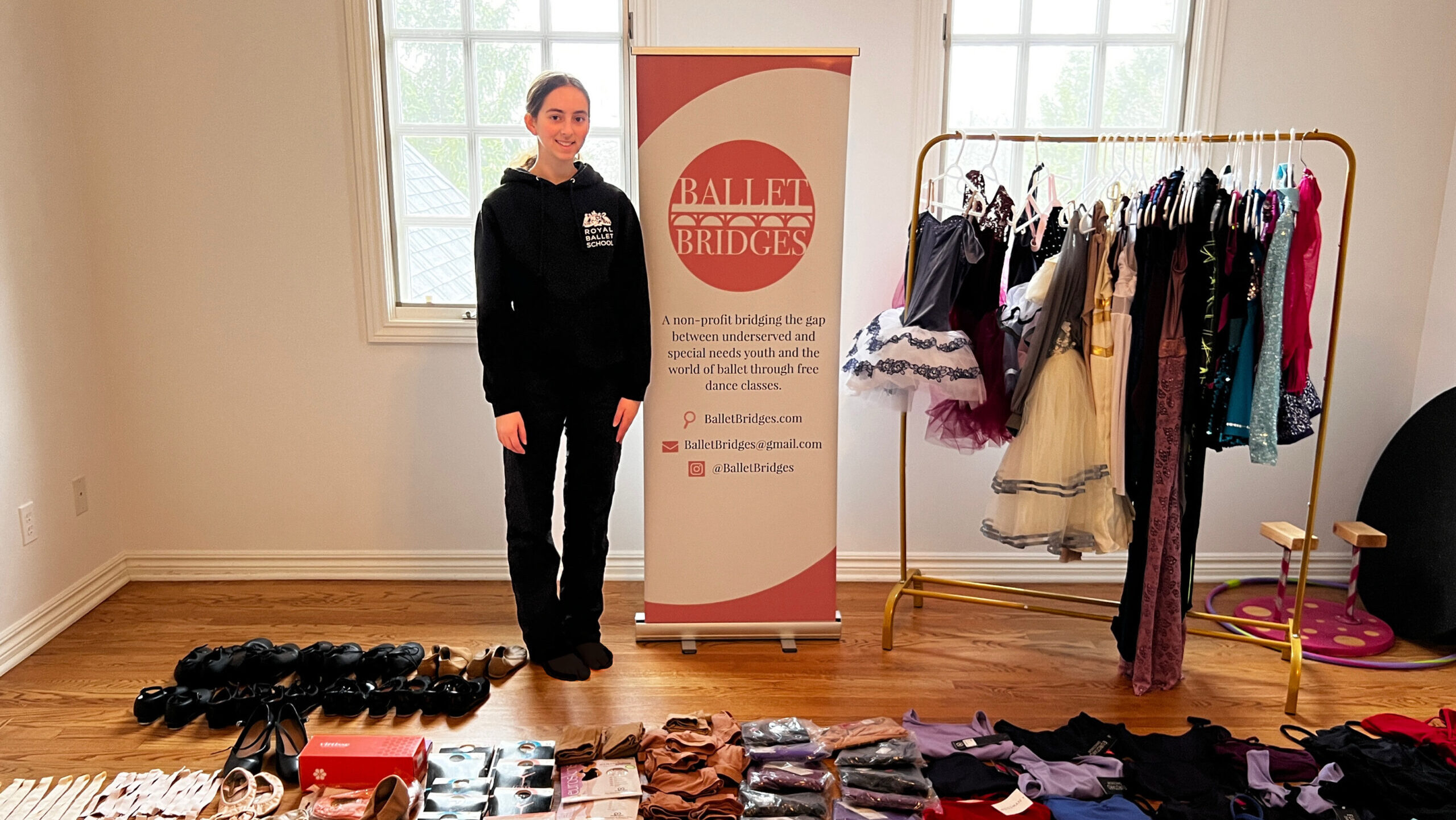 A tenage female ballet student in a black sweatshirt and sweatpants stands next to a vertical banner that says Ballet Bridges. Dance clothing and shoes are in organized piles on the floor in front of her, and a wardrobe rack full of costumes ub to her left.