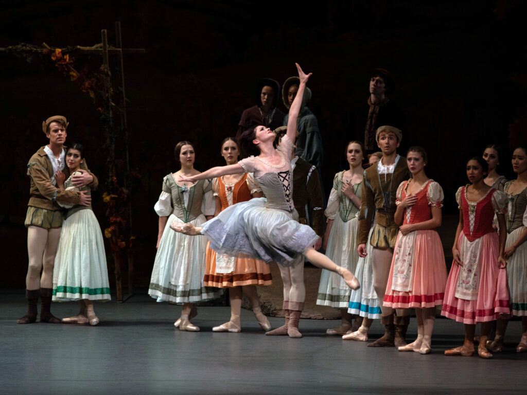 Cassandra Trenary, as Giselle in Act 1, jetés forward widly, reaching up to the sky as she looks past her top hand. Her hair, left down, flies around her head as her blue peasant dress billows. Behind her, corps members dressed as villagers stand and watch with worried expressions.