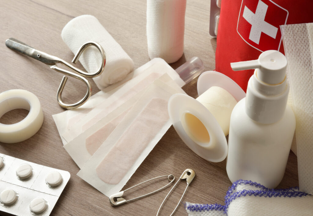 An assortment of first aid items are pictured including tape, medication, bandages, gauze, scissors, and safety pins.