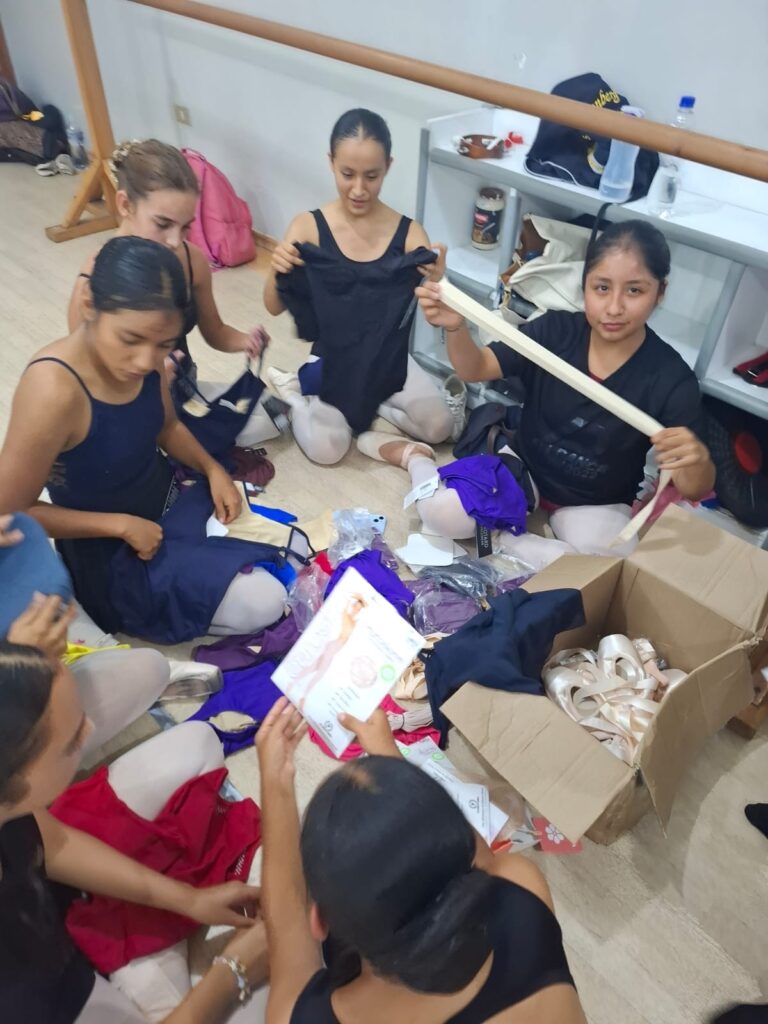 A group of teenage ballet students in practice clothes sit on a dance studio floor and open a box full of pointe shoes, dancewear, and tights.