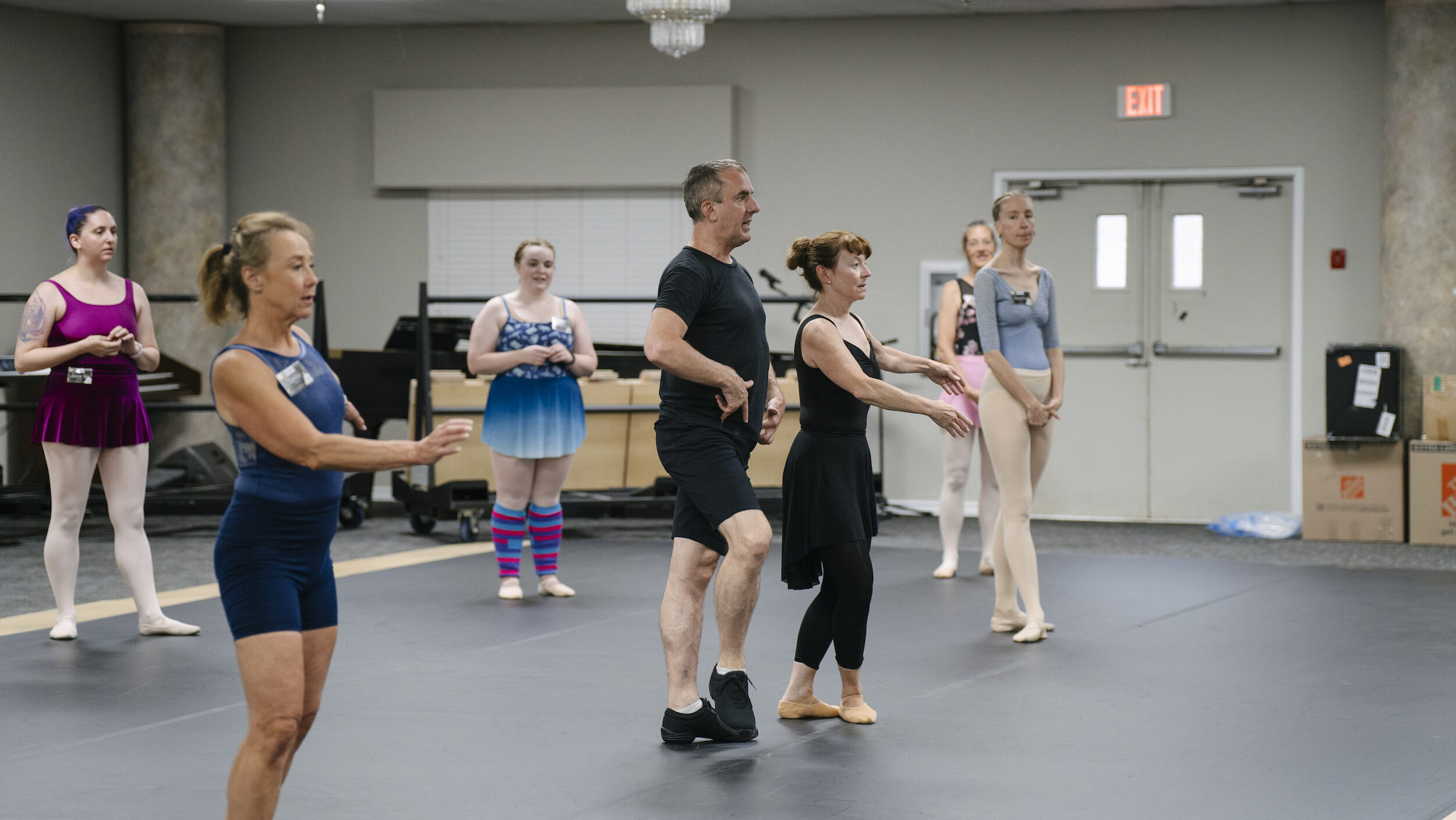 A group of adult dancers in practice clothes listen to a teacher give a correction. Four dancers watch in the background, while the male teacher points to his hip in correcting the female adult student. Another woman marks the exercise to his right.