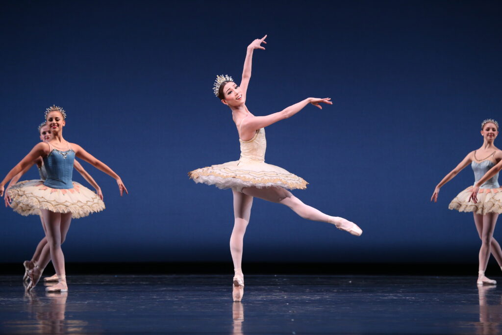 Onstage in front of a blue backdrop, Soo Youn Cho performs in a light gold tutu, standing on pointe in profile with one leg extended devant, her arms in allongé devant as she twists her torso front. Lines of corps dancers pose on either side of her.
