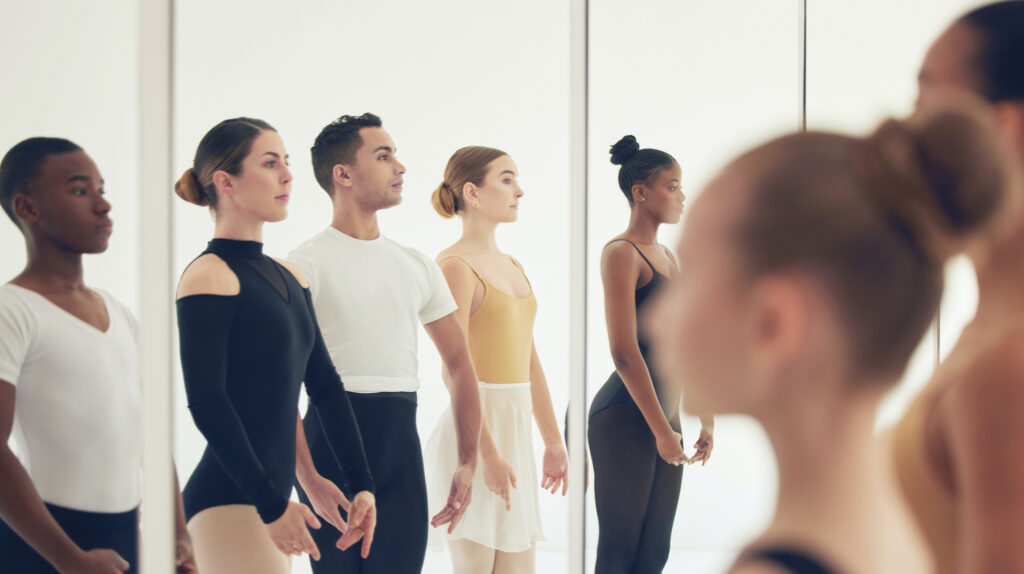 Five ballet dancers in practice clothes stand close to a mirror in fifth position and look at their reflections.
