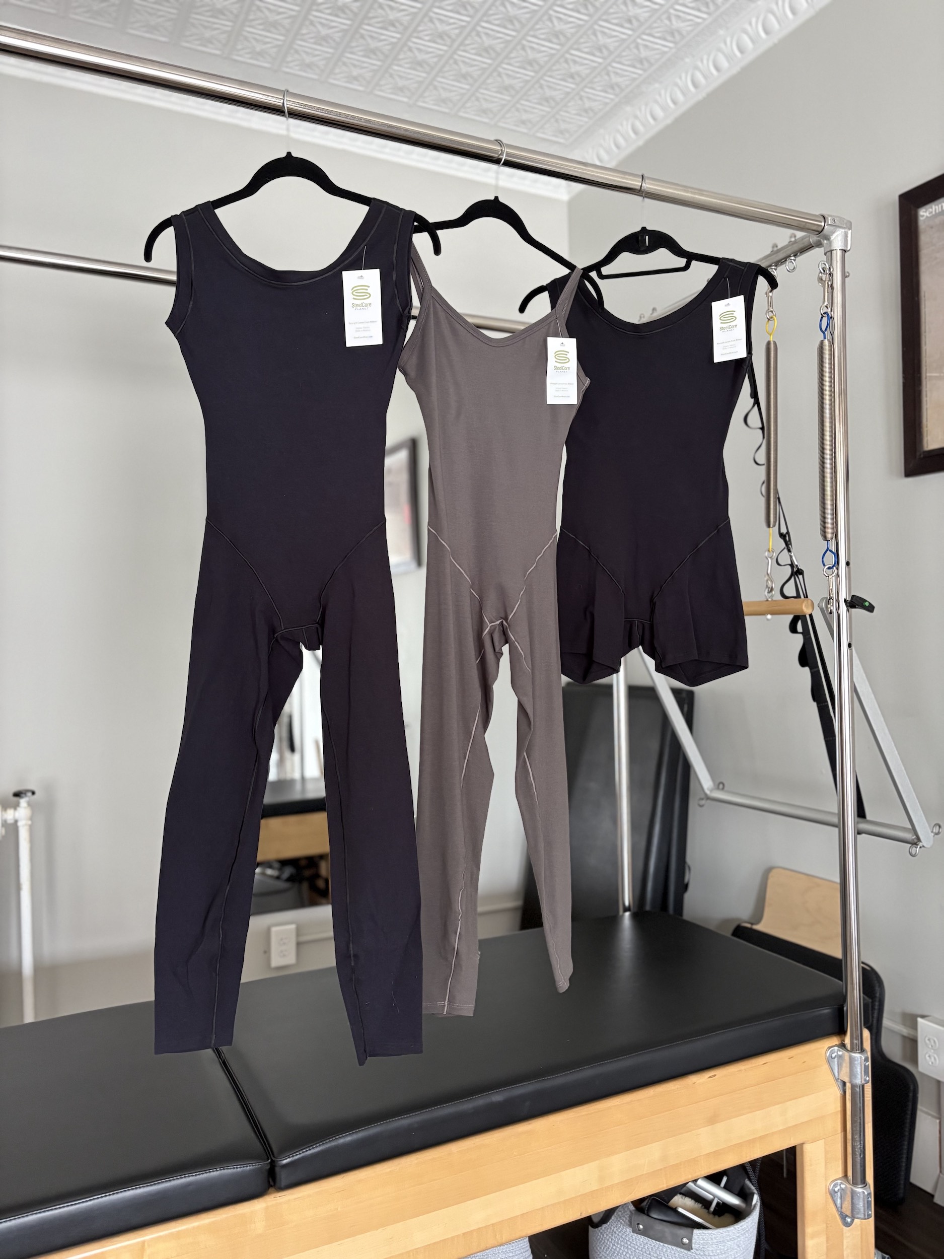 Three pieces of dancewear with tags on them—a black unitard, a gray unitard, and a black biketard— hang on hangers across the bar of a Pilates Cadillac reformer.