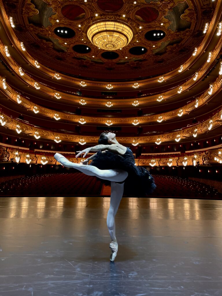 Madeline Woo poses in a majestic attitude derriere en pointe onstage in a grand theater. She arches back over her leg, arms draped backward, and she wears a black pancake tutu.