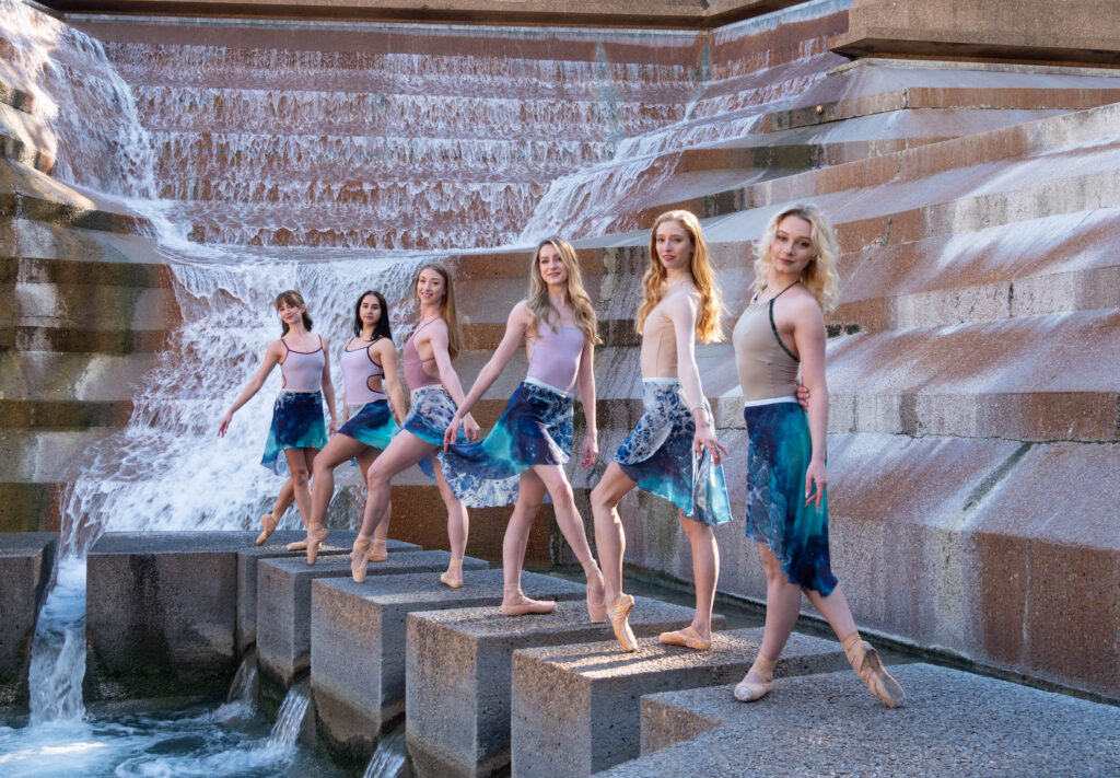 Six ballet dancers in neutral colored leotards, multi-colored blue ballet skirts, and pointe shoes pose in front of a large, modern water fountain.