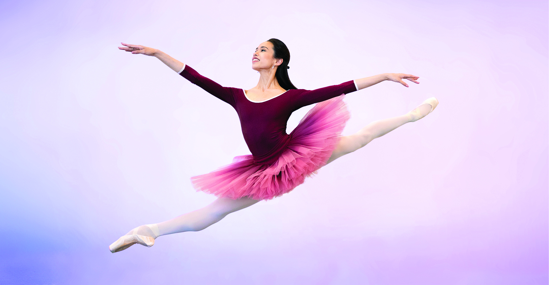 Léa Fleytoux wears a maroon, long sleeve leotard and an ombre maroon/pink tutu while doing a sissone jump in front of a purple backdrop.