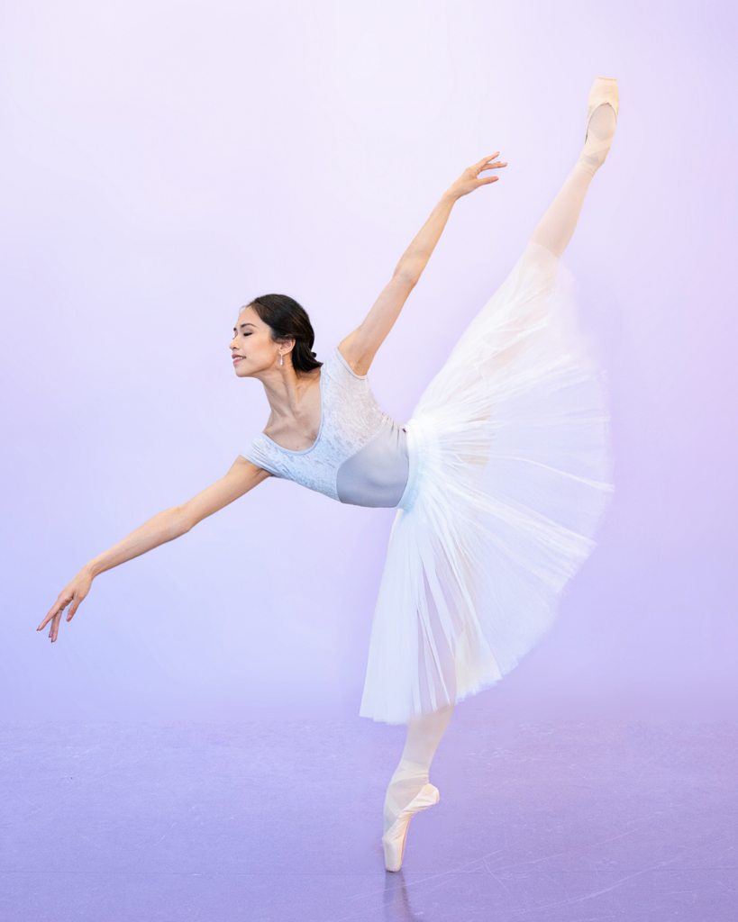 Léa Fleytoux extends her left leg in a first arabesque penché en pointe. She wears a light gray, cap sleeve leotard with velvet lining at the top and a white, romantic tutu.