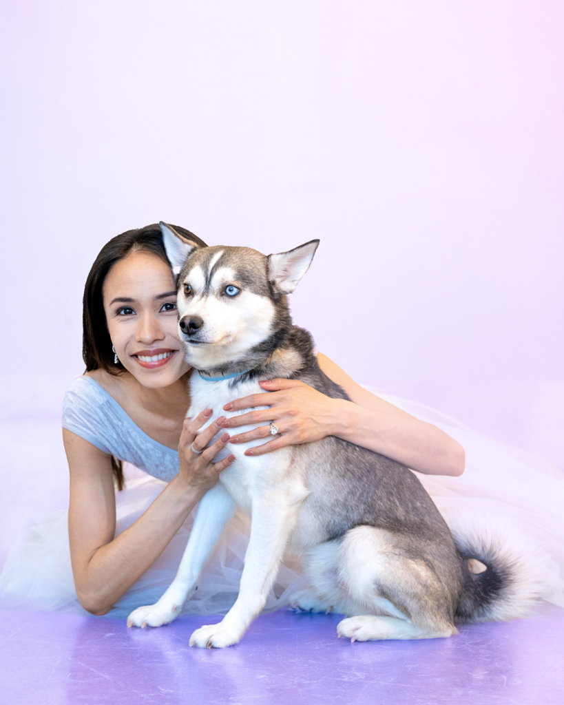 Léa Fleytoux sits on the floor and poses with her dog. The dog, a white and gray husky, stares past the camera while Léa smiles. She wears a light gray, cap sleeve leotard with velvet lining at the top and a white, romantic tutu.