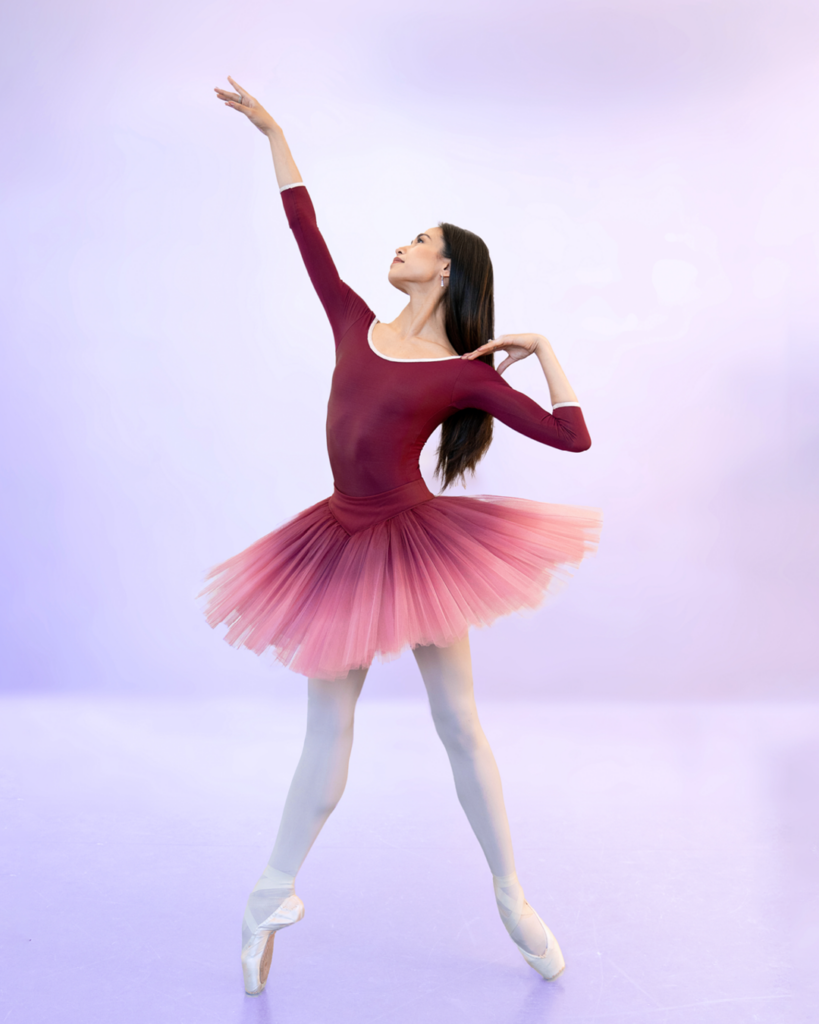 Léa Fleytoux poses in a maroon, long sleeve leotard and an ombre, maroon/pink tutu. She stands en pointe in an echappé with her left hand on her shoulder and right arm extended.