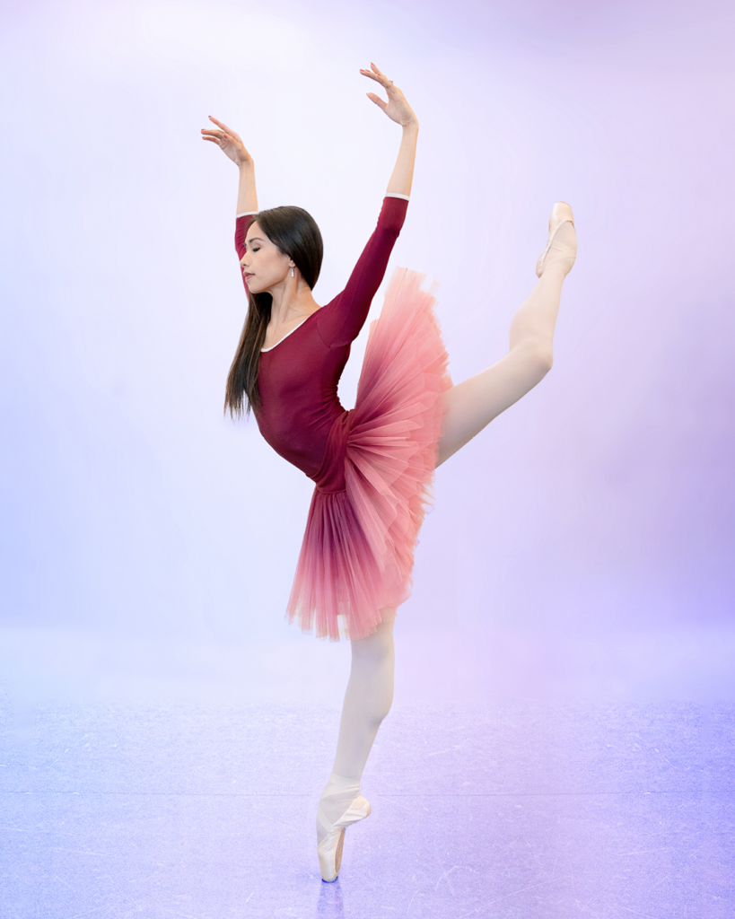 Léa Fleytoux wears a maroon, long sleeve leotard and an ombre maroon/pink tutu against a purple backdrop. She is in a back attitude en pointe with her left leg in the air. Her arms are in an open fifth position and she gazes toward the floor.