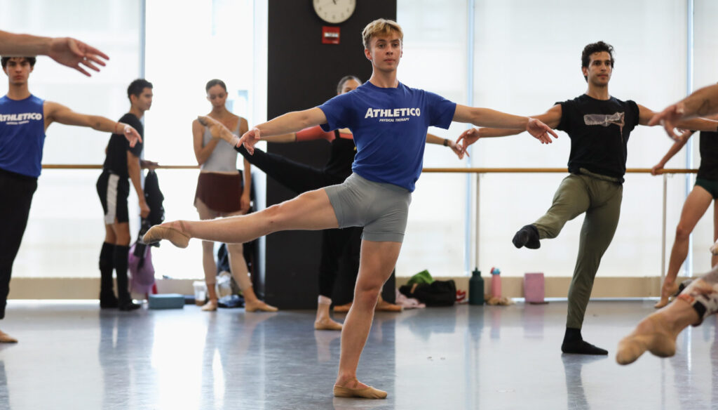 In a large ballet studio, dancers do an adagio combination with legs held in a la seconde, arms in second position. One dancer in the middle wears a blue shirt that says "Athletico Physical Therapy."