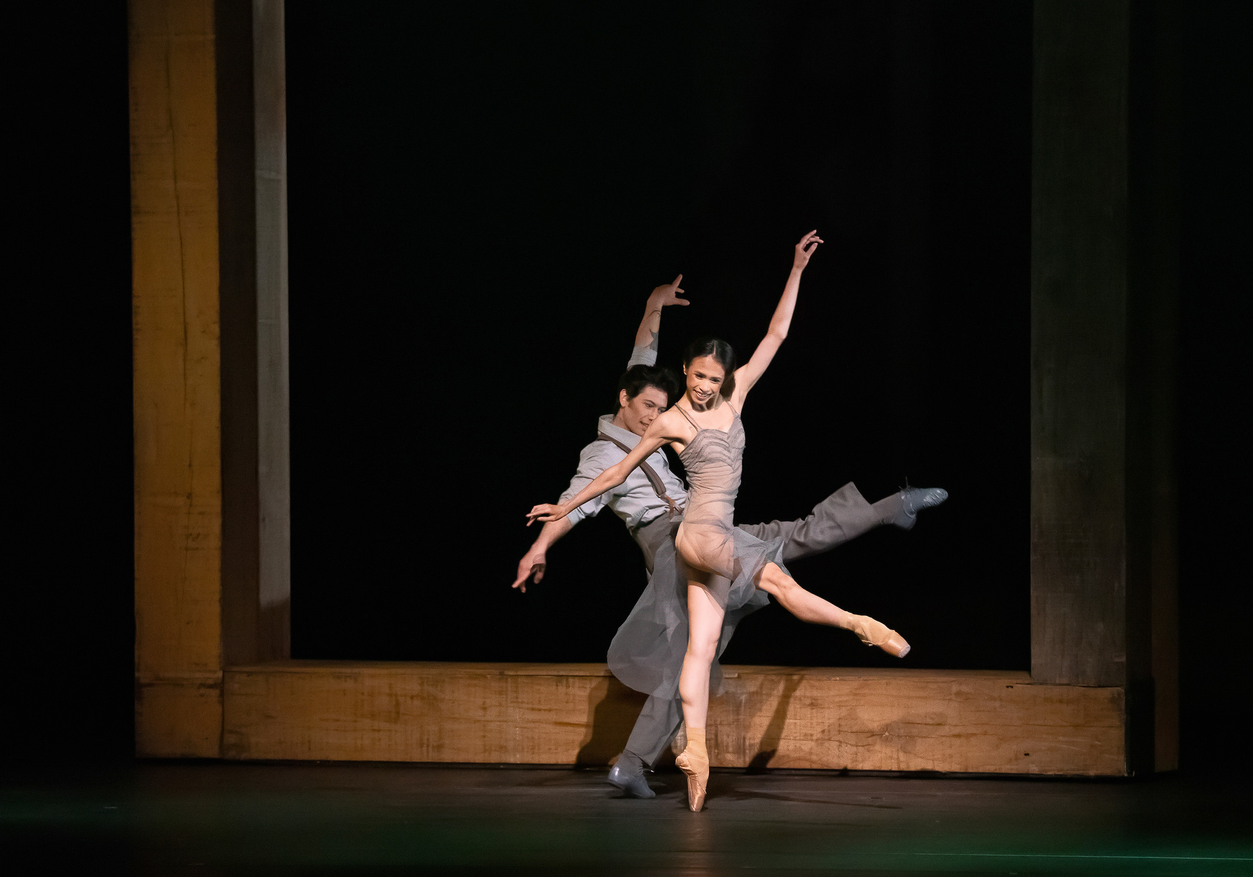 Léa Fleytoux dances en pointe with her right leg extended in a degagé to the front. Her left arm is raised as she smiles at the audience. She wears a flowy gray dress. Herman Cornejo dances behind her in the same degage position on flat. He wears a blue shirt and trousers with suspenders.