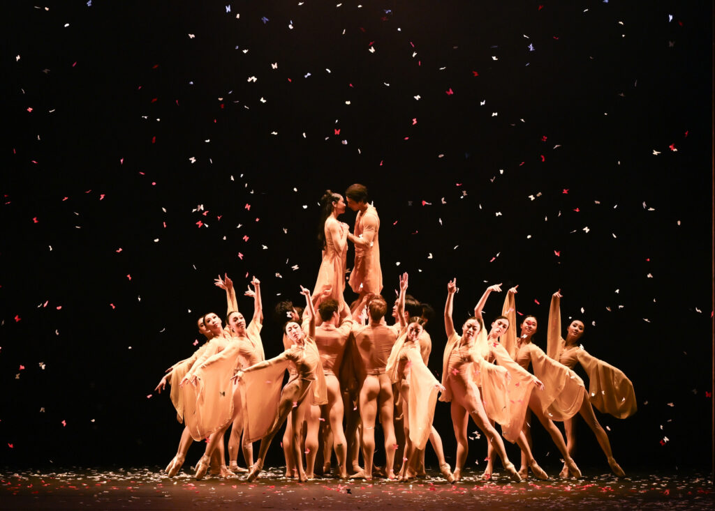 A group of corps dancers dressed in costumes with long, wing-like sleeves pose in a circle surrounding two principal dancers who embrace as they're lifted by a smaller group below. Confetti falls from above.