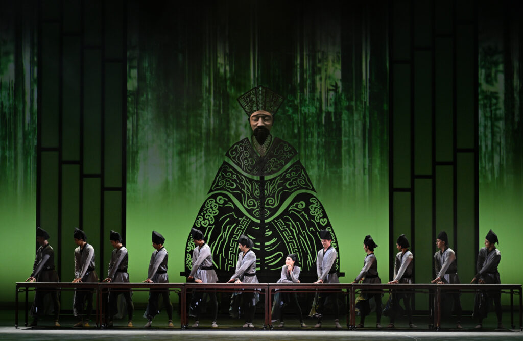 A line of dancers, dressed as students, pose along a long table in front of a backdrop with bright and dark green. A large set piece of a man in formal dress stands center stage behind them.