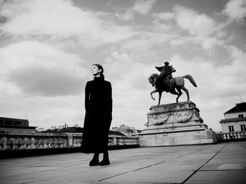 Alessandra Ferria, wearing a black turtleneck and long dark coat, stands in a public square in front of a statue of a man on a horse. She looks towards her right and off into the distance.