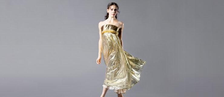 Clara Ruf Maldonado wears a speakly green-gold dress with an empire waist. She stands on a gray backdrop in fourth position croisé on pointe with her arms at her sides.