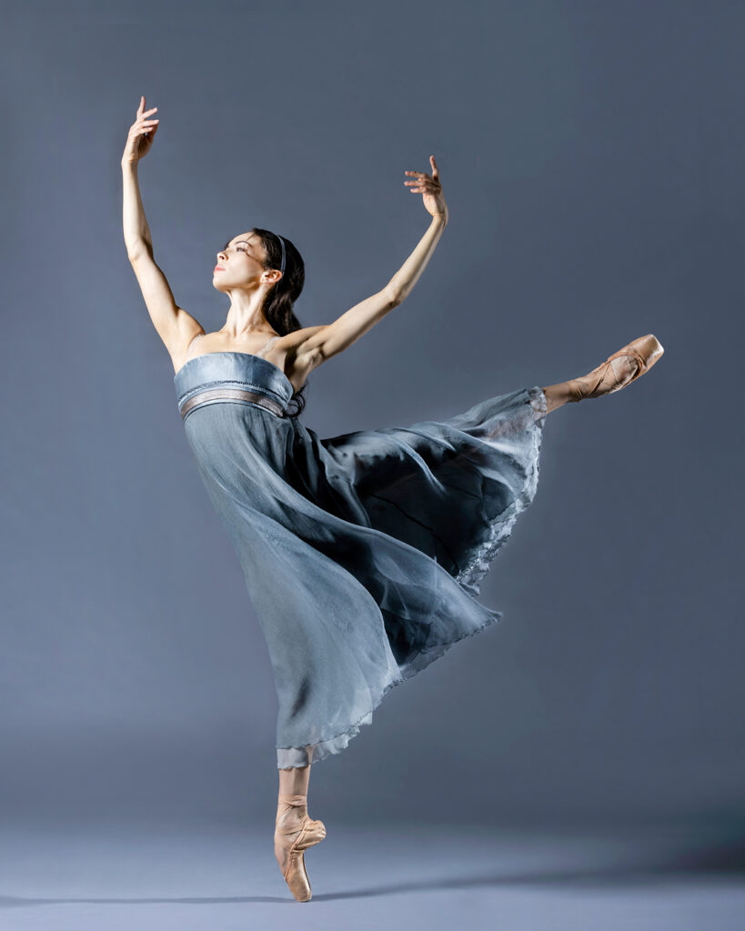 Clara Ruf Maldonado wears a blue empire waist dress and does an arabesque on pointe in frotn of a dusty blue backdrop. She raises her curved arms in a wide fifth position.