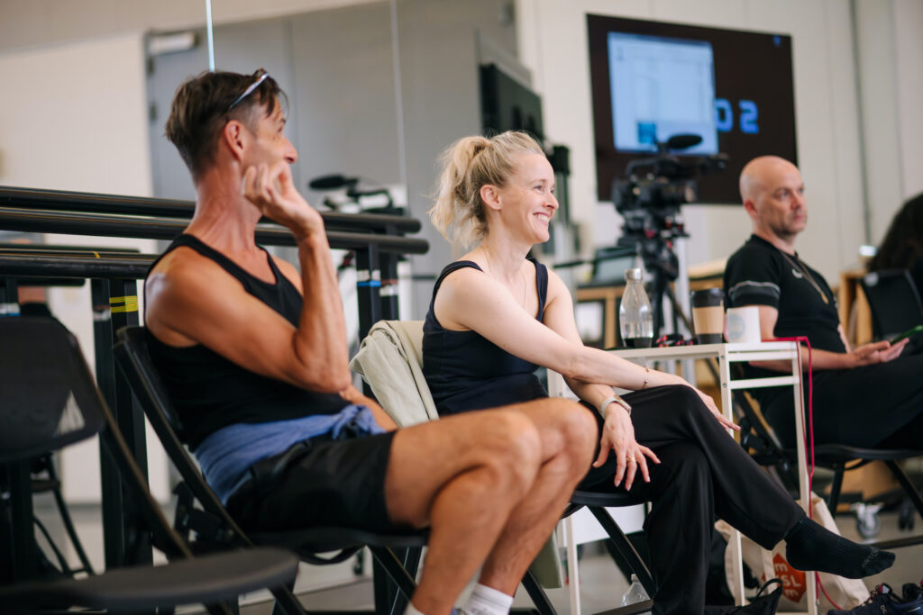 James and Sophie sit in a studio watching rehearsal. James hold his right hand up while speaking to the dancers and Sophie smiles.