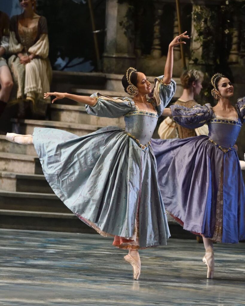 Courtney wears a long, blue dress with ruffled sleeves and an ornate head piece while she does a pique arabesque. Her left arm is raised and she looks toward stage left. Another dancer piques alongside her.