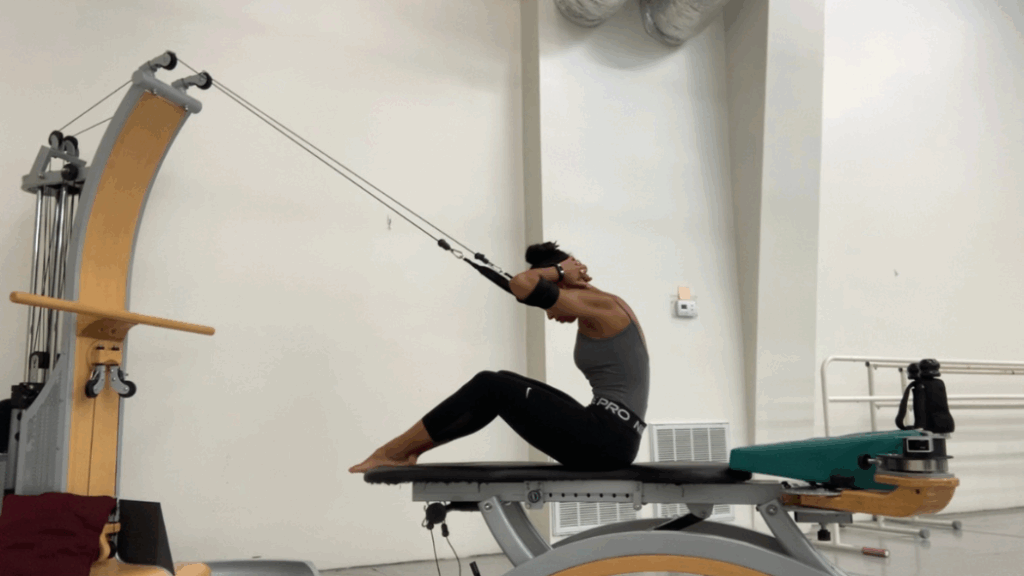 Felecia Baker sits on a Gyrotonic machine with her legs bent, and she curls over her knees with her arms bent, elbows pointing outwards, as her hands rest on the back of her head. She looks down at her lap. Two Gyrotonic straps are looped around her arms, creating resistance.