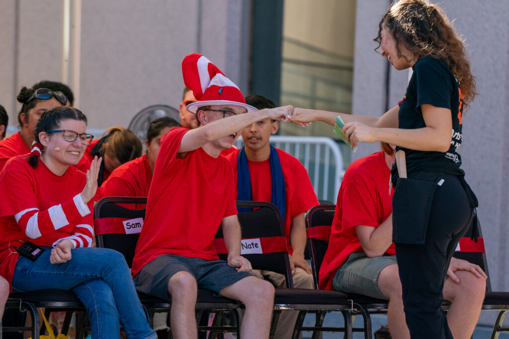 Aneesa fist bumps a youth performer of Studio D's musical theater program. She wears all black and smiles at the performer who is amongst the group of students in red, Suessical costumes.