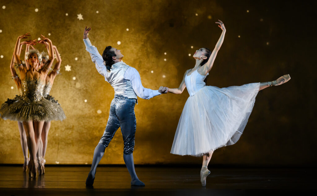 Yu does a pique arabesque with her left leg extended and left arm in fifth, holding onto Lachlan with her right hand. She wears a white bodice and romantic tutu. Lachlan holds her hand with his right, his left arm is in fifth position, and his left leg in a tendu back. He wears gray trousers and tights with a white shirt and silver vest. A group of dancers in ornate silver tutus stands in a line in fifth position on stage right.